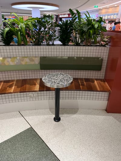 This image captures a bright, modern interior public space, identified as Indooroopilly, Australia, likely a shopping center or food court. The scene is well-lit, suggesting daytime.

The foreground features various seating options and a small table. A long planter box filled with lush, dark green foliage, including plants resembling ZZ plants, Dracaena, and Monstera, runs horizontally, acting as a divider. Behind the plants, there is a section of built-in bench seating. Parts of the bench are upholstered in a solid dark green fabric, while another segment is a lighter green with a subtle geometric pattern. Interspersed with the upholstered sections are segments of warm-toned wooden seating.

A small, circular table with a textured, silvery-gray surface, possibly terrazzo or an aggregate material, stands on a black cylindrical base in front of the seating. The wall directly behind the seating and table is covered in white square tiles with dark grout, forming a grid pattern. A white double electrical outlet is visible on this tiled wall. The floor is a light speckled concrete or terrazzo, transitioning to a darker green speckled section, suggesting different zones or pathways.

In the background, beyond the planters and seating, several people are visible, some blurred, moving or standing in what appears to be a bustling commercial area. Storefronts are discernible, with the prominent red and yellow "McDonald's" sign clearly visible. A portion of the "KFC" logo is also faintly visible below the McDonald's sign. A large, circular, illuminated light fixture hangs from the ceiling in the upper-left background, contributing to the bright ambiance.

The overall atmosphere is functional and modern, characteristic of a contemporary shopping mall's common area designed for resting or dining.