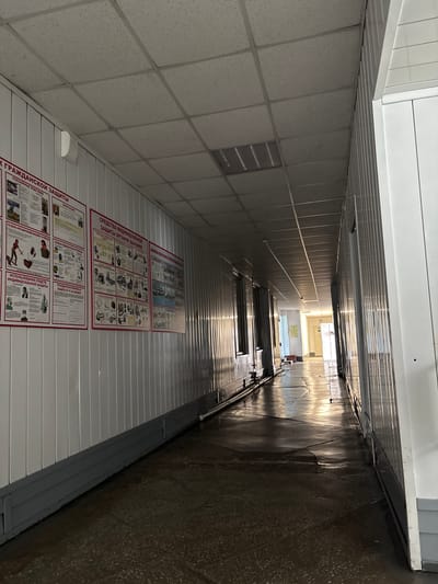 The image depicts a long, narrow interior corridor. The walls are lined with white vertical panels, and the floor appears to be concrete with a dark, slightly reflective surface, showing some signs of wear and cracking. There are overhead fluorescent light fixtures in a suspended ceiling grid. On the left wall, several large informational posters are mounted. These posters contain text and illustrations, likely related to safety or emergency procedures, with titles visible in Russian such as "ГРАЖДАНСКОЙ ЗАЩИТЫ" (Civil Defense) and "СРЕДСТВА ИНДИВИДУАЛЬНОЙ ЗАЩИТЫ" (Personal Protective Equipment). The corridor extends into the distance, with light at the far end suggesting an exit or another area. Sunlight or bright artificial light is reflecting off the walls and floor from the right side, creating a strong glare and casting shadows. There are no people visible in the corridor. The overall impression is of a functional, utilitarian space, possibly within an industrial or institutional building. This scene is located in the city of Belovo, Russia.