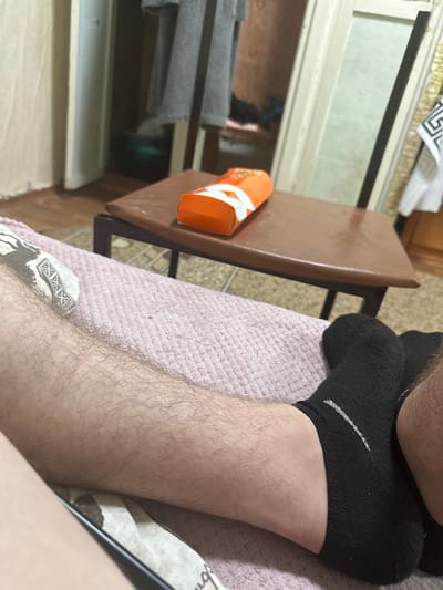 The image shows a close-up of a person's leg and foot, wearing black socks, resting on a pink blanket. In the background, a brown table with an orange box on it is visible, along with a doorway and part of a room. The scene appears to be indoors. There is no indication of the specific location being Corlăteni, Moldova.