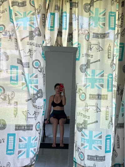 A vertical, medium-shot photograph displays a woman reflected in a mirror, flanked by curtains with a London-themed print, against a tile wall, within an interior space. The curtains, displaying Union Jack flags, London landmarks (Big Ben, Tower Bridge), London phone boxes, motorcycles, guitars, and the word "LONDON," drape on either side of the mirror. The woman in the reflection holds a red cellphone, presumably taking the photograph. She is wearing a black sports bra and patterned shorts. Behind her in the reflection is a dark object, possibly a chair, and a tile wall. The location and city cannot be determined solely from the image.