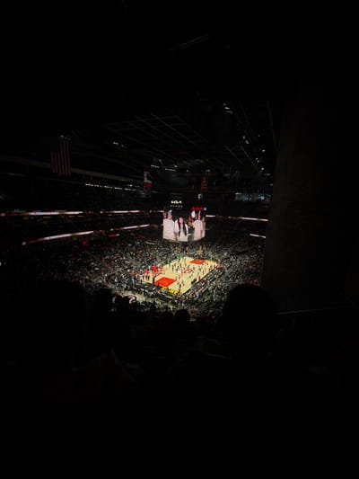The image depicts a basketball game taking place inside an arena. The view is from a high vantage point, likely the upper levels of seating. The basketball court is brightly lit, while the surrounding audience and arena structure are shrouded in darkness. The audience is densely packed, suggesting a well-attended game. A large screen hangs above the court displaying images, and there are American and Canadian flags. The location is inferred to be in Atlanta, United States of America, given the arena’s red color scheme and design which is consistent with the State Farm Arena.