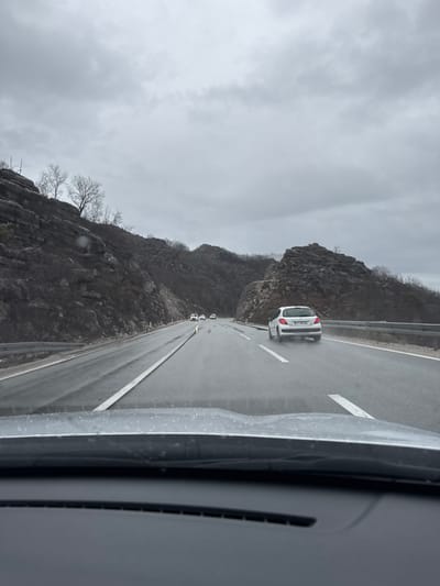 The image captures a wet, multi-lane highway in Gornji Ulići, Montenegro, viewed from the interior of a vehicle. The sky is uniformly overcast and grey. The road surface is visibly damp, with reflections. A white hatchback, identified by a partially visible Montenegrin license plate, is positioned in the right lane ahead of the camera vehicle, its rear tires generating a fine spray of water. Several other vehicles with illuminated headlights are present further down the road, receding into the distance. The highway is flanked on both sides by rocky, dark-colored hills, sparsely covered with leafless trees and dark brush. A metal guardrail runs along the right edge of the highway.