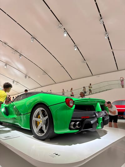 The image shows the interior of the Museo Enzo Ferrari in Modena, Italy, featuring a vibrant green Ferrari LaFerrari on display. The car is positioned on a white platform with spotlights illuminating it from the arched ceiling. Several people are observing the car and the surrounding exhibits. In the background, a red Ferrari is also visible. The architecture of the museum is modern, with a curved ceiling and a grand staircase leading to an upper level where more visitors are present. The overall scene captures the atmosphere of a car museum, highlighting the iconic vehicle and the visitor experience.