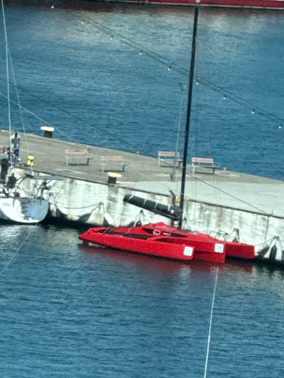 An aerial shot shows a harbor with several boats docked at a pier. In the foreground, a bright red, futuristic-looking catamaran with two hulls is tethered to the dock. The catamaran has a black boom extending from its mast and appears to be empty. To the left, a white sailboat is docked. The pier itself is made of concrete and has several bollards, two benches, and what appear to be life preservers or buoys. The water is a deep blue, and the sky is overcast. The scene is of a working harbor, likely in Gdynia, Poland, a city known for its maritime activity and shipbuilding industry.