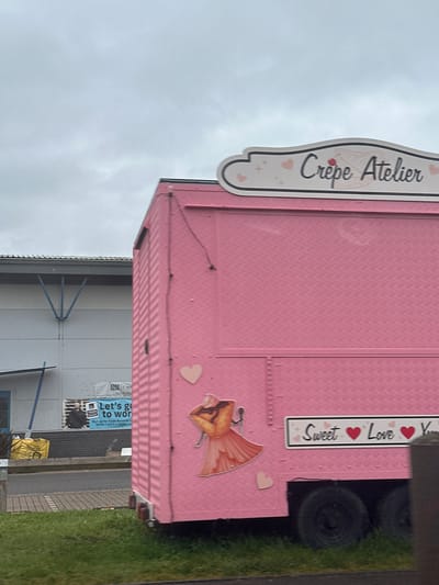 A pink commercial food trailer, branded "Crêpe Atelier," is positioned on a grassy verge next to a paved area under an overcast sky. The trailer's roof features a white sign with "Crêpe Atelier" in black script, accompanied by small pink hearts and a crepe graphic. The trailer's textured pink side is decorated with a large orange dress decal, several heart shapes (pink and white), and a white rectangular sign displaying "Sweet ❤️ Love ❤️ Y[...]" with red heart icons. Two black wheels are partially visible beneath the trailer on the right.

In the background, a large, light-grey commercial building with blue 'V'-shaped structural supports is visible. A blue banner on the building's brick base is partially obscured, displaying "Let's g[o] to wor[k]," "Sign up for Trade Rewards t[o]," and "OPEN 7 DAYS A WEEK," alongside a male figure and the word "TRADE." A yellow bulk bag and a blue pole are visible near the building. A dark post partially obstructs the lower right portion of the trailer. The paved area separates the grassy verge from the building.

The location is identified as the vicinity of Selco Builders Warehouse on Paycocke Road, Basildon, United Kingdom.