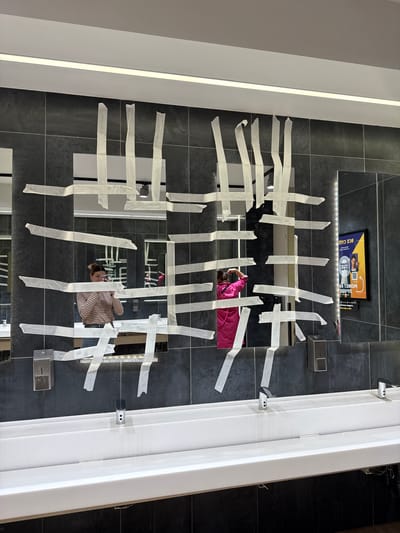 The image, taken from a high-angle perspective, depicts a restroom in Саларьево, Russia. The floor and walls are tiled in a dark grey, stone-like material. A series of square mirrors are mounted on the wall, spaced vertically and horizontally. Each mirror is covered with strips of masking tape arranged in a grid pattern.
Reflected in the mirrors are two individuals. One individual, wearing a pink top, is positioned towards the upper half of the frame. The second individual, wearing a striped shirt, is reflected in the lower half of the mirrors.
A white, rectangular structure, possibly part of the wall or a dividing panel, is present on the right side of the frame. A small advertisement display is visible above the top mirror. Soap or sanitizer dispensers are mounted near the mirrors on the right.