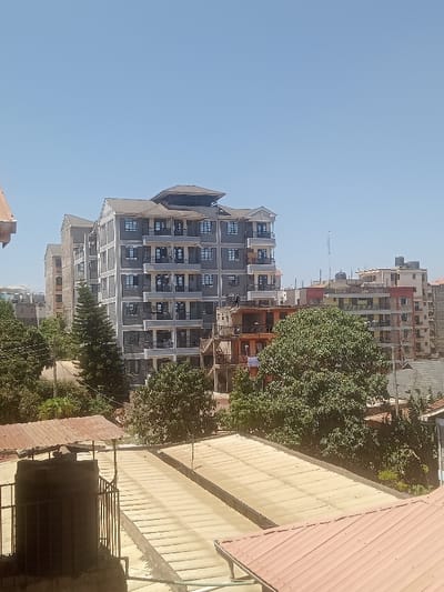 The image depicts an urban landscape under a clear blue sky, likely within Gatongora ward, Kenya. In the midground, a prominent, multi-story apartment building, possibly seven to eight stories high, features a light grey facade with darker grey or black stone accents and multiple balconies with dark railings. Its roof is dark and includes an elevated central section. To its left, another similar, slightly less complete building is visible.
Surrounding the main building are several other structures, appearing to be residential or mixed-use, some in various stages of construction with exposed brickwork or scaffolding. Green trees and dense foliage are interspersed among the buildings, particularly in the midground and lower-left, indicating significant vegetation.
In the immediate foreground, a collection of rooftops is visible. On the left, a corrugated reddish-brown metallic roof partially obscures a dark water storage tank enclosed by metal bars. Adjacent to this, two long, flat, light-colored roofs, possibly concrete or light-colored metal, extend towards the center. Another reddish-brown corrugated roof is visible on the lower right.
No individuals or explicit actions are discernible within the frame. The overall impression is one of ongoing urban development and residential density in a sunny climate.