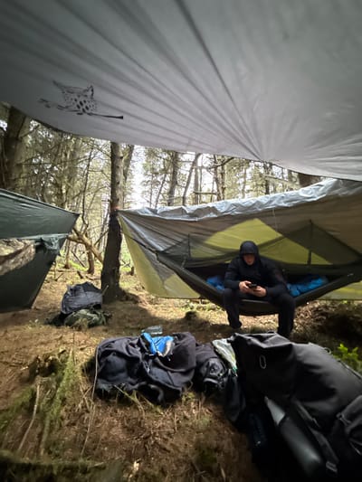 A man is sitting in a hammock in Macclesfield Forest. He is wearing a black hoodie and black pants. He is looking down at his phone. The hammock is suspended between two trees. The man is sitting under a tarp, which is stretched between the trees. There are two bags on the ground in front of the hammock. The forest floor is covered in green moss and brown leaves. The image was taken from a low angle, looking up at the man and the hammock. The coordinates indicate a location in Macclesfield Forest, United Kingdom. The forest is known for its tall trees and dense foliage. The weather is cloudy and overcast. The scene is tranquil and peaceful.