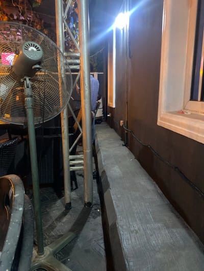 The image depicts an illuminated outdoor or semi-outdoor commercial area at night in Igbessa, Nigeria. In the foreground on the left, a large, industrial-style standing fan with a light green/grey finish and a metal cage is positioned on a four-pronged base. Behind the fan, a silver-colored metal truss structure with horizontal bracing extends vertically. To the left of the truss, dark, woven chairs are visible, and a seated individual, seen from the back, wears a blue and white checkered shirt. Overhead, subtle illumination highlights what appears to be decorative foliage.
On the right side of the image, a narrow, elevated concrete walkway extends into the background, bordered by a dark brown building wall. The walkway surface is textured. Two white-framed windows are set into the wall, emitting bright white light that causes significant lens flare. Dark electrical cables are secured along the lower section of the wall, running parallel to the walkway, and a white electrical outlet is visible. Shadows are cast by the fan and truss onto the patterned dark ground beneath.