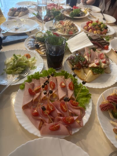 The image depicts a table laden with various dishes, suggesting a gathering or meal. The setting appears to be an indoor dining area, with a pale, textured tablecloth covering the surface. On the table, there are several white plates, some containing food items, and others empty or partially cleared.
In the foreground, a white plate showcases rolled slices of ham, each garnished with a cherry tomato and a small black olive skewered on a toothpick. These are arranged on a bed of lettuce. Adjacent to this, another white plate holds several open-faced sandwiches or bruschetta, topped with ingredients such as chopped tomatoes, olives, and herbs. Further back, a glass of dark liquid, likely a beverage, sits on the table.
Other plates visible include one with a salad, another with what appears to be sliced meat and possibly some herbs, and a plate with fish and lemon. The presence of multiple forks and glasses suggests a meal or a party.
The lighting in the image is soft and diffused, indicating it may be taken indoors during the daytime or with artificial lighting. The overall impression is one of a social occasion centered around food. The image does not provide any specific clues that would allow for a definitive identification of the location as Zelenaya Polyana, Russia, nor are there any people clearly visible or identifiable within the frame.