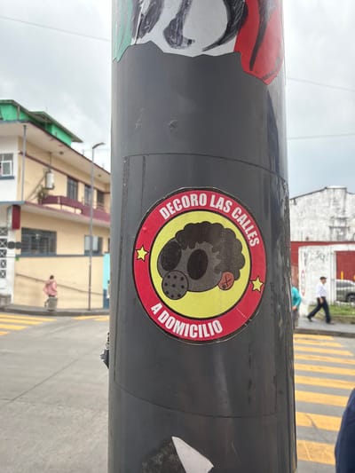 A gray pole displays stickers. One depicts a stylized dark figure in a yellow circle bordered by red, with yellow stars. Text circles the figure: "DECORO LAS CALLES A DOMICILIO." Additional stickers show parts of a face. The background features a cityscape, including a multi-story building, street, and people.