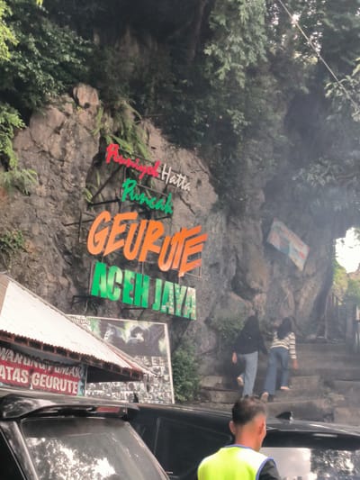 The image depicts an outdoor scene featuring a rocky cliff face adorned with signage and vegetation. A prominent sign in vibrant orange and green lettering reads "GEURUTE ACEH JAYA," with smaller red lettering above it that appears to say "Rumah" and "Puncak". To the left, a corrugated metal roof extends from a building or structure, with additional signage below it that is partially obscured but seems to indicate "WARUNG" and "ATAS GEURUTE". Behind this structure, a large billboard displays images of what appear to be monkeys. A stone staircase ascends the cliff to the right, with two individuals, one wearing a black top and jeans, and the other a striped shirt and jeans, walking up the stairs. In the foreground, the rear of a dark-colored vehicle is visible, and a person wearing a bright yellow vest over a black shirt is seen from behind, facing away from the camera. The overall environment suggests a tourist destination or natural attraction, likely in Meudheun, Indonesia, given the signage. The lighting is somewhat overcast, with a slight haze present in the air.