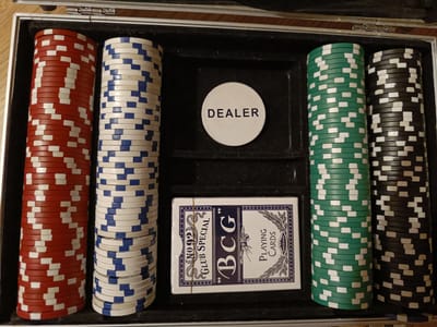 The image shows an open poker chip set with four stacks of poker chips and a deck of playing cards. The chips are colored red, blue and white, green and white, and black and white. A circular "DEALER" chip is placed in the center of the set. The playing cards are in a blue and white box with the brand name "BCG" and "PLAYING CARDS" visible. The setting appears to be indoors, with the poker set resting on a wooden surface. The image provides no information about the time of day, weather, or any emotions. The location context "Hovden, Norway" is provided but not visually evident in the image.