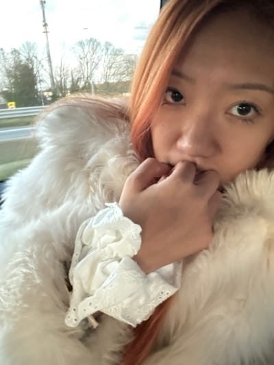 A woman with reddish-blonde hair, dark eyes, and freckles is partially visible. She is wearing a fluffy white fur coat and a white blouse with lace detailing. Her hand is near her face. A window is in the background, showing a blurred landscape with trees and a utility pole. The image is rotated counter-clockwise, giving a tilted perspective.