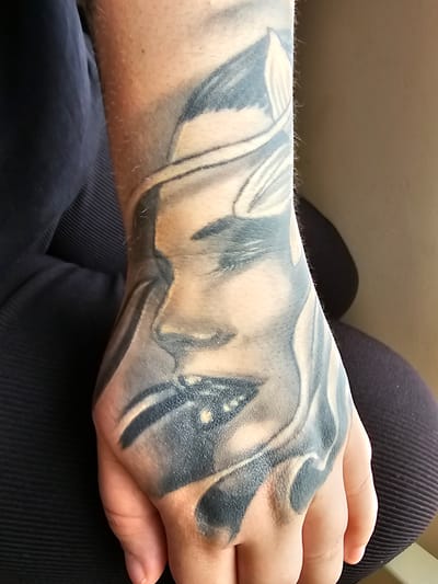 An adult arm and hand with a black and gray tattoo are resting on a lap. The tattoo covers the entire hand and extends up the forearm. The person is wearing dark, possibly black, trousers. The background is blurry and indistinct. There is no indication of the location of Anderlecht, Belgium.