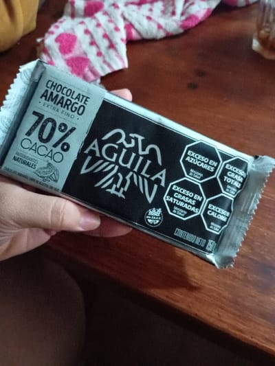 A person is holding a wrapped bar of Aguila Negra 70% Cacao dark chocolate, "Chocolate Amargo Extra Fino," in their right hand. It is located on a wooden surface, possibly a table. The wrapper is black with white lettering and a hexagonal symbol warning of excessive sugars, saturated fats, and calories. The person is wearing a pink and white patterned shirt. The chocolate bar is likely located in a private residence, possibly in El Talar, Argentina, a suburb of Buenos Aires, based on the provided coordinates. There are other objects in the background, but they are blurred and unrecognizable.