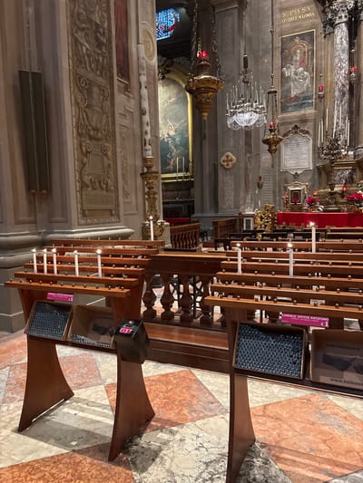 The image displays the interior of the Cattedrale di San Giorgio Martire in Ferrara, Italy, viewed from the nave towards the chancel and altar area. The scene is illuminated by ambient artificial lighting and flickering candlelight.
In the foreground, two freestanding, dark-stained wooden votive candle stands are positioned on a geometrically patterned marble floor composed of reddish-brown, white, and light-brown tiles. Each stand features horizontal wooden bars with metal pegs designed to hold candles. The left stand holds seven lit white votive candles, while the right stand holds two lit white votive candles. Below the candle racks, both stands contain cardboard boxes filled with unlit, dark grey/black pellet-form "anti-smoke" votive candles. A pink sign on the right stand reads "CANDELE VOTIVE ANTIFUMO €1" (Anti-smoke votive candles €1). Between the stands, a dark columnar device with a red digital display showing "01" is present, likely a payment terminal.
The midground features a dark wooden balustrade with turned spindles, separating the foreground from rows of dark wooden pews. Beyond the pews, an ornate side altar, partially draped in red fabric with small lit candles, is visible.
The background reveals the cathedral's architectural details. On the left wall, ornate bas-relief panels with figures and decorative motifs are visible, alongside a tall, dark grey speaker unit. A large, decorated paschal candle on a golden stand is positioned here, with a small lit candle at its base. High above, a vibrant stained-glass window and a large religious painting depicting multiple figures are visible. To the right, a rectangular painting of "S. PIVS V. PONTIFEX MAXIMVS" (Saint Pius V, Supreme Pontiff) hangs above a marble inscribed plaque. Multiple ornate chandeliers, including golden fixtures with red lights and a crystal chandelier, suspend from the high ceiling. An ornate altar area on the far right is adorned with red cloths, gold ornamentation, poinsettia plants, and several small lit candles. No individuals are present in the scene.