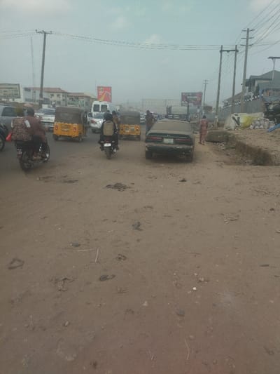 The image shows a street in Way, Nigeria, at coordinates 9.029 latitude and 7.584 longitude. There are three yellow, three-wheeled motorbikes parked in the middle of the street, two on the left and one on the right, with a few people standing around them. Further down the street, a dark-colored car is parked, and a person is walking away from the car. The street is covered in a light-brown dust, and there are several power lines running above the street. Buildings are visible in the background, and there are several cars driving on the street in the distance.
