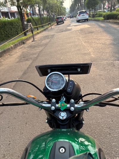 The image shows a first-person perspective from a green motorcycle riding on a street. The motorcycle's handlebars and dashboard, including the speedometer, are visible in the foreground. The speedometer reads approximately 35 km/h, with an odometer reading of 059225 km. The road is paved with asphalt and appears to be in moderate condition with some patching. Trees line the side of the road, and there are buildings in the background. Several cars are present on the road, indicating moderate traffic. A yellow caution tape is visible along the side of the road, supported by black posts. The lighting suggests it is late afternoon or early evening, with sunlight casting long shadows. The location is identified as Pakulonan Barat, Indonesia.