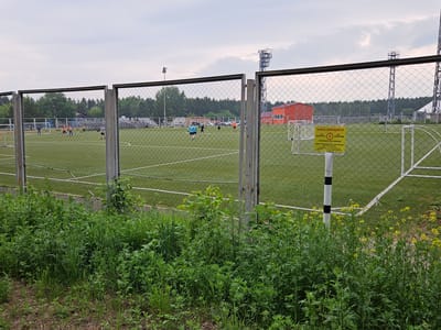 The image depicts a fenced-in artificial turf soccer field in Sosnovoborsk, Russia. Several individuals are playing soccer on the field. A bright orange building is visible in the background, along with a telecommunications tower and stadium lighting. A yellow sign with black text, warning of a 2-meter safety zone around a cable, is posted on a pole near the fence. The foreground is overgrown with tall green grass and some yellow wildflowers. The sky is overcast.