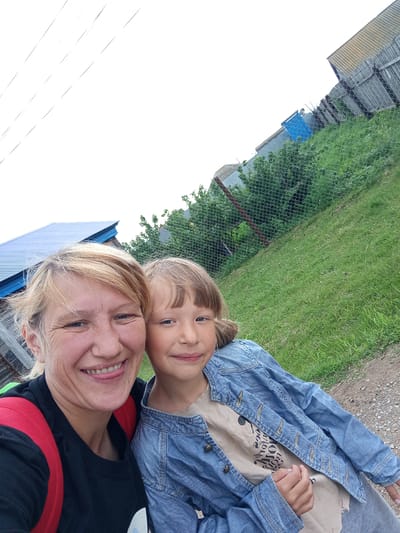 Two people, an adult female and a child, are taking a selfie outdoors in what appears to be a rural area. The adult female, on the left, has blonde hair and is wearing a black t-shirt with a red backpack. She is smiling and sticking out her tongue. The child, on the right, has light brown hair and is wearing a denim jacket over a light-colored t-shirt. They are both looking at the camera. In the background, there is a green grassy hill with a chain-link fence running across it. Behind the fence, there are several rustic wooden structures, some with corrugated metal roofs, and a blue-roofed building. The sky is overcast. The location is identified as Buraly, Russia.