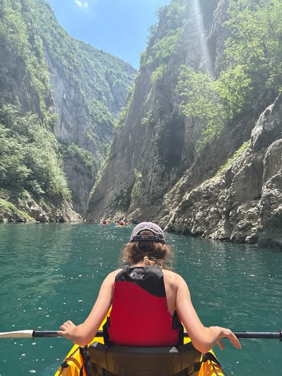 A person is kayaking on a bright, sunny day in a picturesque canyon. The water is a vibrant turquoise, reflecting the sunlight and creating shimmering patterns on its surface. Steep, rugged cliffs rise dramatically on either side of the river, covered in lush green vegetation. The cliffs are textured and imposing, with evident layers of rock.

The kayaker, viewed from behind, is wearing a red and black life vest and a grey baseball cap. Their arm is extended, holding a paddle that rests on the yellow kayak. The person appears to be enjoying the activity, with their posture suggesting a relaxed and engaged posture.

In the distance, other kayakers can be seen navigating the waterway, forming a small flotilla. The sunlight casts rays through the canyon, illuminating parts of the scene and creating a slightly hazy effect in some areas. The clear blue sky is visible above the towering cliffs.

The overall scene evokes a sense of adventure and immersion in nature. The scale of the canyon, the vibrant colors of the water and vegetation, and the presence of multiple kayakers suggest a popular recreational spot for outdoor activities. The time of day is clearly daytime, likely late morning or early afternoon, given the strong sunlight. There is no visible text in the image.