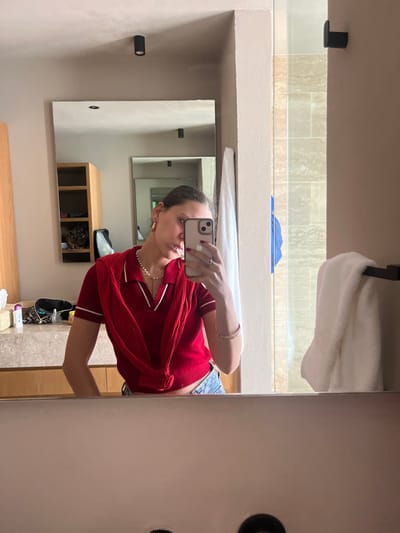 The image displays a female individual taking a mirror selfie in a modern bathroom. The person is reflected from the waist up, holding a light-colored smartphone with a dual-lens camera system in her right hand, which partially obscures her lower face. She has dark hair pulled back, gold hoop earrings, a white pearl necklace with a small gold pendant, and a gold bangle bracelet on her left wrist. Her fingernails are painted red. She wears a red short-sleeved polo shirt with white trim on the collar and sleeves, and a matching red knitted garment is tied around her shoulders and chest. The top portion of blue jeans is visible below her shirt.
The bathroom features a large rectangular wall-mounted mirror above a light-colored stone or marble countertop. On the left side of the counter, various small containers and a black bag are visible. To the left of the reflected image, an open-shelf wooden cabinet is present. The walls are painted a neutral light beige or taupe. Two black, cylindrical spotlight fixtures are mounted on the ceiling. To the right, a shower enclosure with clear glass panels and light-toned, possibly travertine, tiled walls is visible, with a blue towel hanging inside. A white folded towel is draped over a black towel bar mounted on the wall adjacent to the shower. The overall environment suggests a modern, well-appointed bathroom, potentially within a residence or hotel in Valle de Bravo, Mexico; however, the exact location or specific address within the city cannot be ascertained from the image content.