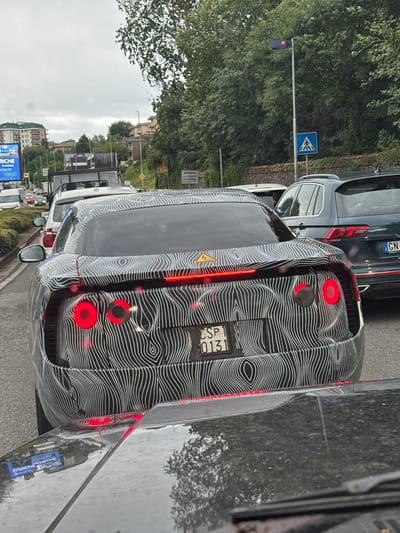 This is a medium shot of the rear of a black car with a white wavy pattern, parked on a street. The car has its red brake lights on. The license plate is visible and reads "CSP 0131". The car is in traffic, with other vehicles visible in front and behind it. The scene is set in Lipomo, Italy, indicated by a road sign with Italian text and a pedestrian crossing sign. The weather appears to be overcast, and the ground is wet, suggesting recent rain. Trees line the street, and buildings are visible in the background. The overall mood is mundane, capturing a typical moment in urban traffic.