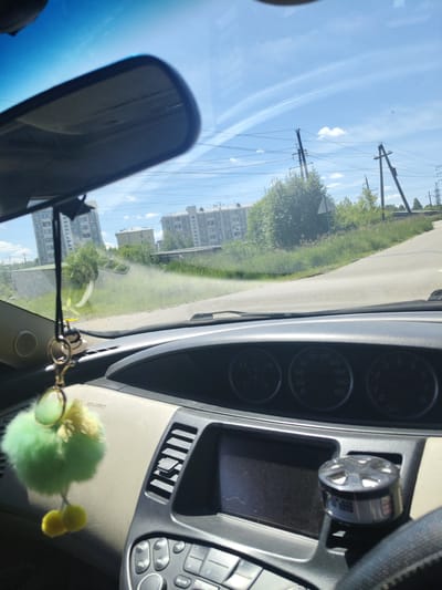 The image shows the interior of a car with a dashboard and windshield. Outside the car, a road curves to the right, bordered by grass and trees. In the background, apartment buildings and utility poles are visible under a blue sky with white clouds. A fluffy green keychain hangs from the rearview mirror. The dashboard includes a screen, air vents, and gauges.