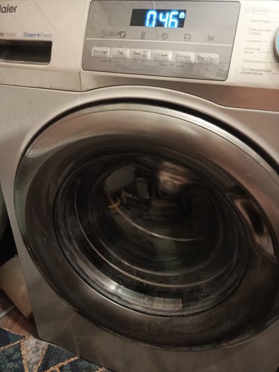 The image shows the control panel and drum of a Haier washing machine. The control panel has a digital display showing "0 46" and various buttons with Russian labels such as "Отл. старт", "Пар", "Темп.", "Отжим", "Полоск.", and "Старт/Пауза". On the right side of the panel are options for different wash cycles like "Хлопок", "Синтетика", "Смешанная стирка", "Гигиена", "Детские вещи", "Ежедневная", and "Дистанционное управление". The washing machine drum is visible, appearing dark and metallic. The surroundings suggest an indoor setting, possibly a laundry room, with a rug partially visible in the foreground. There is no information in the image that indicates the location is the city of Meget, Russia.