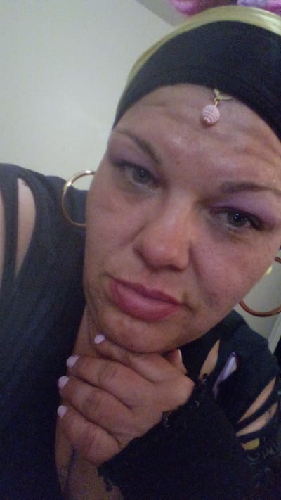 A close-up selfie depicts a woman with light skin, wearing a dark-colored head covering adorned with a small, round, pink ornament dangling from the center of her forehead. She is wearing large hoop earrings and has mauve-colored eye shadow. Her fingernails are painted a light pink. Her hand is positioned under her chin. The environment is nondescript, with a plain wall visible in the background. There is no indication of this picture's location in Tulsa, United States of America.