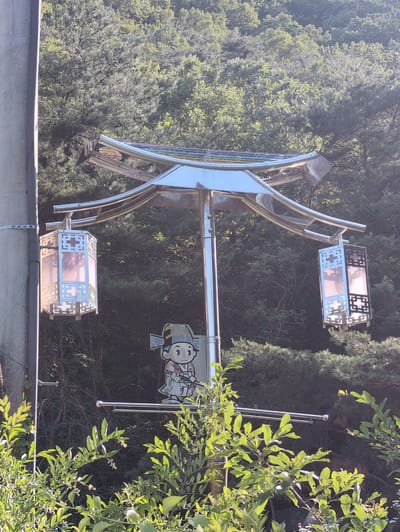 In Godang-ri, South Korea, a modern, highly reflective metallic street light or decorative pole stands against a backdrop of dense green foliage. The structure features a central vertical pole supporting a stylized, double-tiered curved canopy reminiscent of traditional Korean architectural roofs. Two rectangular lanterns, suspended from either side of the main pole, display metallic frames with intricate geometric cutout patterns and translucent, light-diffusing panels. Below the main lighting elements, a cutout figure of a cartoon boy is attached to the pole. The character wears traditional Korean attire, including a striped hat and a white robe with colored accents, and holds a slender stick or brush. A concrete utility pole is partially visible on the far left. The scene is illuminated by natural daylight, highlighting the metallic surfaces, with lush green trees and undergrowth forming the background and foreground.