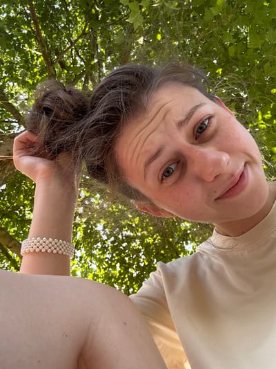 This is a selfie taken from a low angle, looking up towards the subject and the sky. The subject is a person with fair skin, gray-blue eyes, and dark brown hair pulled back. They are wearing a light-colored t-shirt. Their face is turned towards the camera, and they appear to be smiling or smirking. The background is filled with green leaves and branches of trees, suggesting an outdoor setting during the daytime, likely a sunny day given the bright lighting. The subject is holding their hair with one hand, which is adorned with a pearl bracelet. The image composition, with the subject's head tilted and looking up, creates an intimate and slightly playful perspective. The lighting suggests it is daytime, possibly late afternoon due to the soft light. The presence of trees and the natural light point to a natural outdoor environment, perhaps a park or garden. There is no visible text in the image.