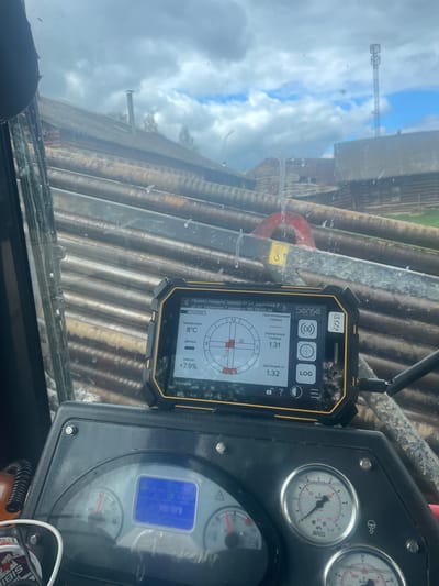 The image shows the interior of a vehicle, presumably a piece of heavy machinery, judging by the dashboard gauges. A smartphone-like device with the brand "Sense" is mounted on the windshield. The device's screen displays data related to a project described as "Проект: поедуги, прокол от ул. заречная 8 до ул. заречная 7, диаметр 110, 09.05.26" (Project: Poedugi, puncture from Zarechnaya St. 8 to Zarechnaya St. 7, diameter 110, 09.05.26). The screen also shows measurements: a predicted depth, current temperature of 8°C, inclination of +7.9%, and a distance of 1.32m.
Through the windshield, a blurred view of pipes or beams and a utility pole is visible. The sky is overcast with a mix of clouds and blue patches. A portion of the dashboard with gauges is visible. The gauges include a pressure gauge with the brand "WIKA" and RPM values. A sticker with the name "Sibis" is on the bottom right.