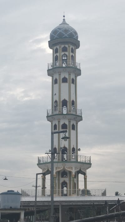 The image captures a tall, white minaret with ornate gold trim, topped by a patterned dome, against a cloudy sky. The minaret has multiple levels, each featuring arched windows and balconies with railings. Loudspeakers are visible on some levels. In the foreground, a metal lamppost with a light fixture is present. Below the minaret, a portion of a white-roofed structure is visible, along with a blue water tank on the left. The scene is set in Bale, Indonesia.