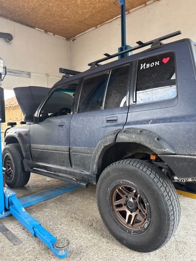 A dark blue SUV is elevated on a blue hydraulic car lift in what appears to be a garage or workshop. The vehicle is dusty, suggesting it has been used off-road or is undergoing maintenance. A rack is mounted on the roof of the SUV. On the rear passenger side window, there is a white sticker of a red heart and the Cyrillic text "Ивон". The vehicle has black fender flares and bronze-colored rims on its tires. The background shows a concrete floor and walls, one of which is clad with Oriented Strand Board (OSB) panels. The lighting suggests it is daytime. The overall scene depicts a vehicle in a state of being worked on, possibly for repairs or modifications, in a practical, unglamorous setting.