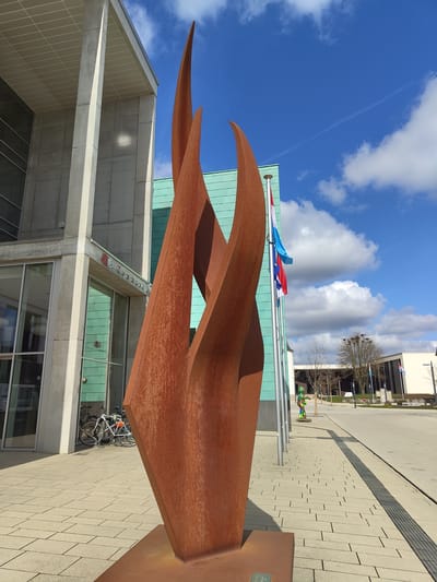 The image depicts an exterior scene in Bertrange, Luxembourg, featuring a large abstract sculpture and modern architecture under a bright blue sky with scattered white clouds and a faint contrail.

In the immediate foreground, a monumental, multi-pronged sculpture crafted from a rust-colored material, likely Corten steel, rises vertically, its forms resembling stylized flames or upward-reaching leaves. To the left of the sculpture, a modern building with a facade of vertical concrete pillars and large reflective glass panels is visible. Above its main entrance, a sign clearly reads "Commune de Bertrange" with a red logo. Two bicycles are parked against the building's facade, secured to a metal bike rack. Adjacent to this building and partially obscured by the sculpture is another structure featuring a teal-colored paneled or tiled facade.

To the right of the main sculpture, a series of silver flagpoles are positioned, one prominently displaying the national flag of Luxembourg, while another flies the flag of the European Union. The ground is a meticulously paved plaza composed of light-colored rectangular pavers, with a dark grey tactile paving strip running longitudinally along a section of the path. Further down the plaza, a colorful, possibly humanoid, statue adorned with green, orange, yellow, and blue patterns stands on the paving. In the background, additional modern buildings with dark windows and a stand of bare deciduous trees are visible, extending into the urban landscape. The scene is illuminated by strong sunlight, casting distinct shadows to the right, and no individuals are actively present.