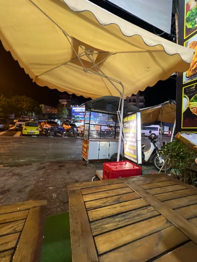 The image depicts a nighttime street scene in Bắc Hà, Vietnam, likely on a paved road or sidewalk. In the foreground, a wooden table is visible, suggesting an outdoor seating area for a food establishment. To the left, a large yellow awning shades a food cart. Behind the cart, several motorbikes are parked, and a yellow car and an orange pickup truck are visible on the street. Further in the background, buildings with illuminated signs and trees line the street. The overall impression is of a bustling, urban environment at night, with indications of commercial activity and food service.