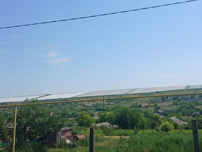 A wide-angle, eye-level shot captures a rural landscape under a clear blue sky, with a power line bisecting the upper portion of the frame. In the middle ground, a long, white, greenhouse-like structure stretches across the horizon, its translucent covering reflecting the sunlight. Below this structure, a village of scattered houses with grey roofs is nestled among rolling green hills and dense trees. In the foreground, a yellow metal pipe runs horizontally, supported by a vertical post, partially obscuring a lush green garden with overgrown vegetation. A weathered wooden fence encloses the area, with a rusty barrel and some debris visible on the left side. The overall scene depicts a tranquil, sun-drenched day in a rural area, likely the city of Semionovca, Moldova.