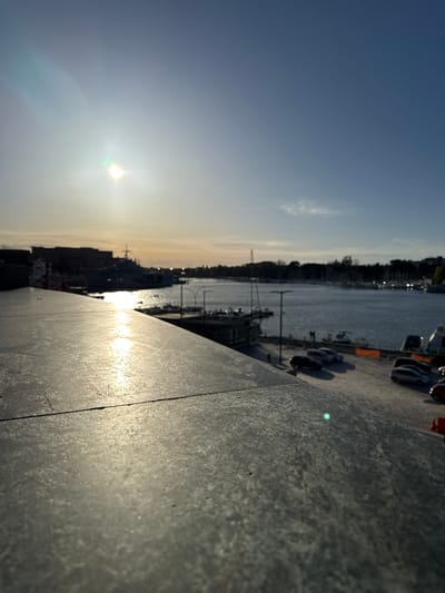 The photograph shows a view from a high vantage point in Brindisi, Italy, oriented to capture the sunset over the harbor.
The composition features a textured, gray stone wall or surface in the foreground, taking up a large portion of the right side of the frame. The sun is low in the sky, emitting a bright glare with prominent lens flare. The harbor is visible in the middle ground, with boats docked along the concrete pier. The water is calm, reflecting the setting sun. Buildings, trees and structures line the shore. The sky transitions from a pale yellow to a deeper blue towards the upper left corner of the image. The perspective is unusual, with a significant tilt.