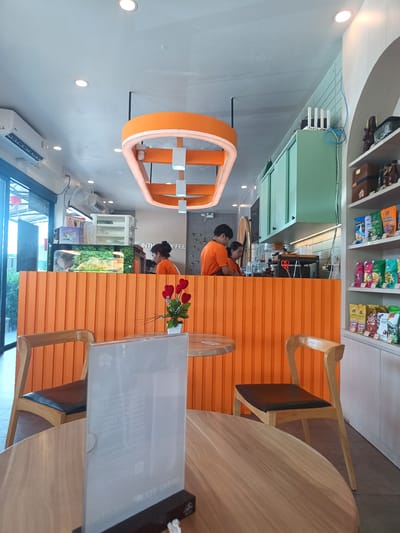 The interior of a coffee shop in Plai Phraya, Thailand, is shown. The scene is captured from the perspective of someone seated at a wooden table in the foreground. On the table is a menu holder, and a vase with red hearts is visible on the orange-colored service counter. Two wooden chairs with black leather seats are arranged around the table. Behind the orange counter, two employees in orange shirts are working. A third employee is also visible behind the counter. The counter has an aquarium. Above the counter are mint-green cabinets and a shelf unit stocked with various snack items. An orange light fixture hangs from the ceiling. A window is visible in the background.