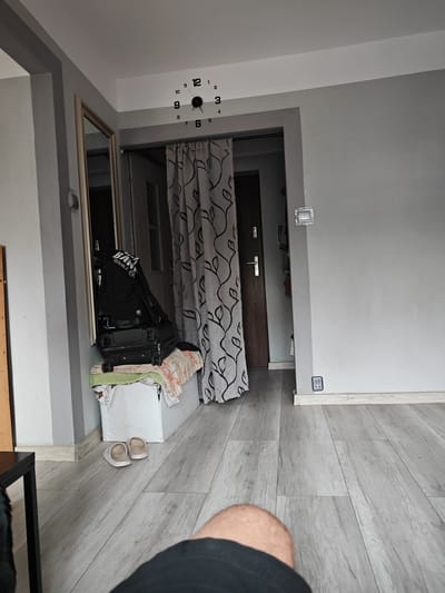 This image shows a living room area with a person's leg in the foreground. The room has light gray walls and a light gray wood-look floor. A large mirror with a silver frame is on the left wall. To the right of the mirror, there is a doorway with a gray patterned curtain. Inside the doorway, a brown door is visible.
On a white storage unit near the doorway, there are two pieces of luggage stacked on top of each other, with a black backpack on top. Several pieces of fabric are draped over the storage unit. A pair of white slippers are on the floor in front of the storage unit.
A wall clock with black numbers is mounted on the ceiling above the doorway. The clock shows the time as approximately 12:17. There are light switches and electrical outlets on the walls. The overall impression is of a lived-in space, possibly a home in Stargard, Poland, given the location context. The lighting suggests it is daytime. There are no people clearly visible other than the leg in the foreground, and no specific event is taking place. The text visible on the backpack reads "BAN TROUBLE".