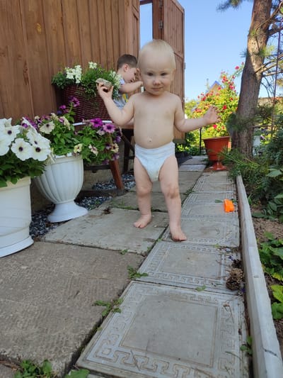 In Sovetsko-Nikol'skoe, Russia, a toddler stands on a stone pathway in a backyard setting. The child, wearing a diaper, is facing the camera with arms outstretched and a slight smile. Behind the toddler, another child is partially visible, reaching into a wicker basket filled with white and purple flowers. The pathway is bordered by potted plants and green foliage, with a wooden fence and a tree visible in the background. The sky is clear and blue.