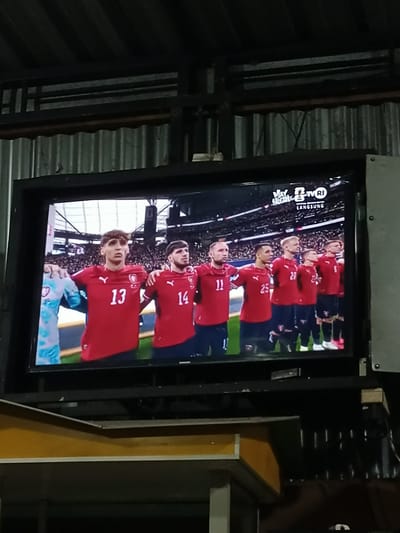 The image captures a television screen displaying a football match, likely a broadcast from TVRI, indicated by the logo in the upper right corner of the screen. The screen shows a line of athletes wearing red jerseys with numbers prominently displayed. The athletes appear to be standing at attention, possibly during a national anthem or a pre-game ceremony. In the foreground, the TV is mounted on a dark metal structure, and the background consists of a corrugated metal roof and some industrial-looking supports. Below the television, a wooden structure is visible, possibly a table or counter. The overall scene suggests an outdoor or semi-outdoor viewing area, perhaps a local eatery or community space in Pulokiton, Indonesia.