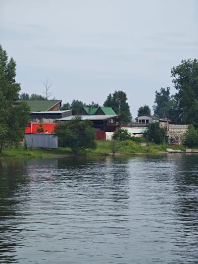 A body of water, likely a river or lake, occupies the foreground of the image. The water's surface is rippled, reflecting the sky and the shoreline. The shoreline in the middle ground is lined with vegetation, including trees and bushes. A cluster of buildings is visible along the shoreline. These appear to be a mix of residential structures and possibly industrial or storage units. One prominent building is a bright red shipping container, next to a corrugated metal structure. Further back, there are several houses with green roofs and wooden siding. A red car is parked on the right side of the shore, near a concrete structure. The sky above is overcast with a pale, grayish-blue hue. The overall impression is a semi-rural or industrial area on the outskirts of a settlement. The location is identified as Ермолаевский Затон, Russia. There are no people visible in the image.