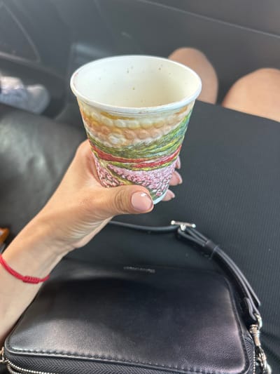 An individual's hand, adorned with a red string bracelet on the wrist and light pink nail polish with a white accent on one finger, holds a paper cup with a patterned design that resembles a landscape with pink flowers and green hills. The cup is positioned in the foreground, slightly tilted. In the background, blurred legs of another person are visible, suggesting they are seated. A black leather bag with silver hardware is placed in the lower part of the frame. The overall scene appears to be within a vehicle. There is no discernible information indicating a specific location in Vodenichane, Bulgaria.
