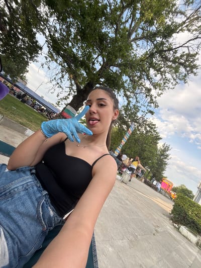 The image depicts a young woman taking a selfie in an outdoor public setting, identified as Kostievo, Bulgaria, by a visible Bulgarian flag. The woman, with dark hair pulled back, is seen from the chest up, angled slightly to the left. She is wearing a black spaghetti-strap top and blue denim jeans, partially visible. On her right hand, she wears a light blue nitrile-style glove and forms a peace sign (V-gesture) with her index and middle fingers, extending towards the camera. Her tongue is slightly protruded. She has a silver necklace with a small pendant and a silver bracelet on her right wrist.
She appears to be sitting or reclining on a dark green patterned fabric, laid on a concrete path or sidewalk. To her left, a grassy area hosts numerous people gathered under blue and white canopies or tents, suggesting a market, fair, or public event. Large trees with green foliage dominate the upper left background and extend across the top of the image.
To her right, a thick tree trunk is visible, around which a Bulgarian flag (white, green, red horizontal stripes) is tied. Further along the concrete path, a vertical structure composed of multiple colorful, rectangular signs is visible. Several individuals are present on the path, including a person in a yellow and black shirt. Stalls displaying various goods, including clothing, are set up along the path, with a bright pink structure visible in the far right midground. The sky above is partly cloudy with patches of blue visible through the tree canopy.