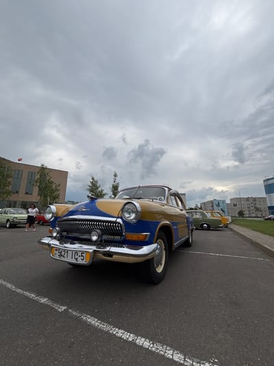 A vintage blue and gold Volga GAZ-21 car is parked on an asphalt surface in front of a beige, modern building with a red and green flag on its facade. The car has a chrome grille, round headlights, and a Belarus license plate that reads "BY 5421 IC-5". A woman in a white shirt and black skirt is walking in the background. The sky is overcast with scattered clouds. Other vintage cars are parked in the background.