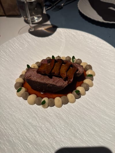 A close-up overhead shot displays a meticulously plated dish of meat with mushrooms and pureed vegetables, served on a textured white plate. The meat, a dark red slice, is topped with thin, glistening mushroom slices. Surrounding the meat is a ring of alternating light beige and grey-brown dollops of puree, each adorned with a tiny green leaf. The purée forms a circular pattern, with some dollops placed on a thin layer of dark reddish sauce. The plate rests on a dark blue surface, with a white textured charger plate visible beneath. A glass of water and silverware are partially visible in the upper left background. The lighting suggests an indoor setting, possibly a restaurant, with a shadow cast on the blue surface. The location is identified as Jianye District, China.