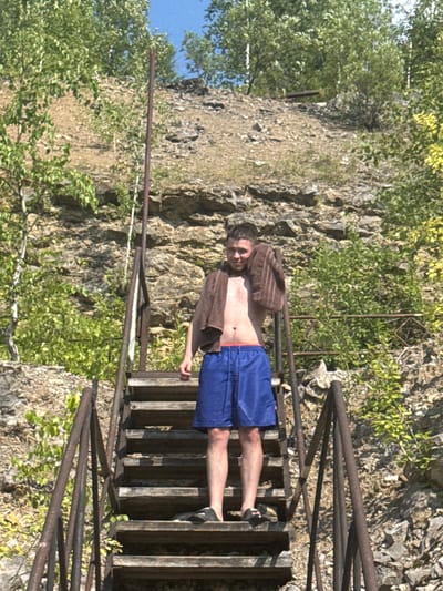 A young Caucasian male stands on a wooden staircase with metal railings, outdoors. He is wearing blue swim trunks and has a brown towel draped over his shoulders. The staircase appears to be in a natural setting with a rocky, sloped embankment covered in vegetation behind him. The sky is visible at the top of the frame, indicating it is daytime. The scene is located in the city of Karyer Izvestnyak, Russia.